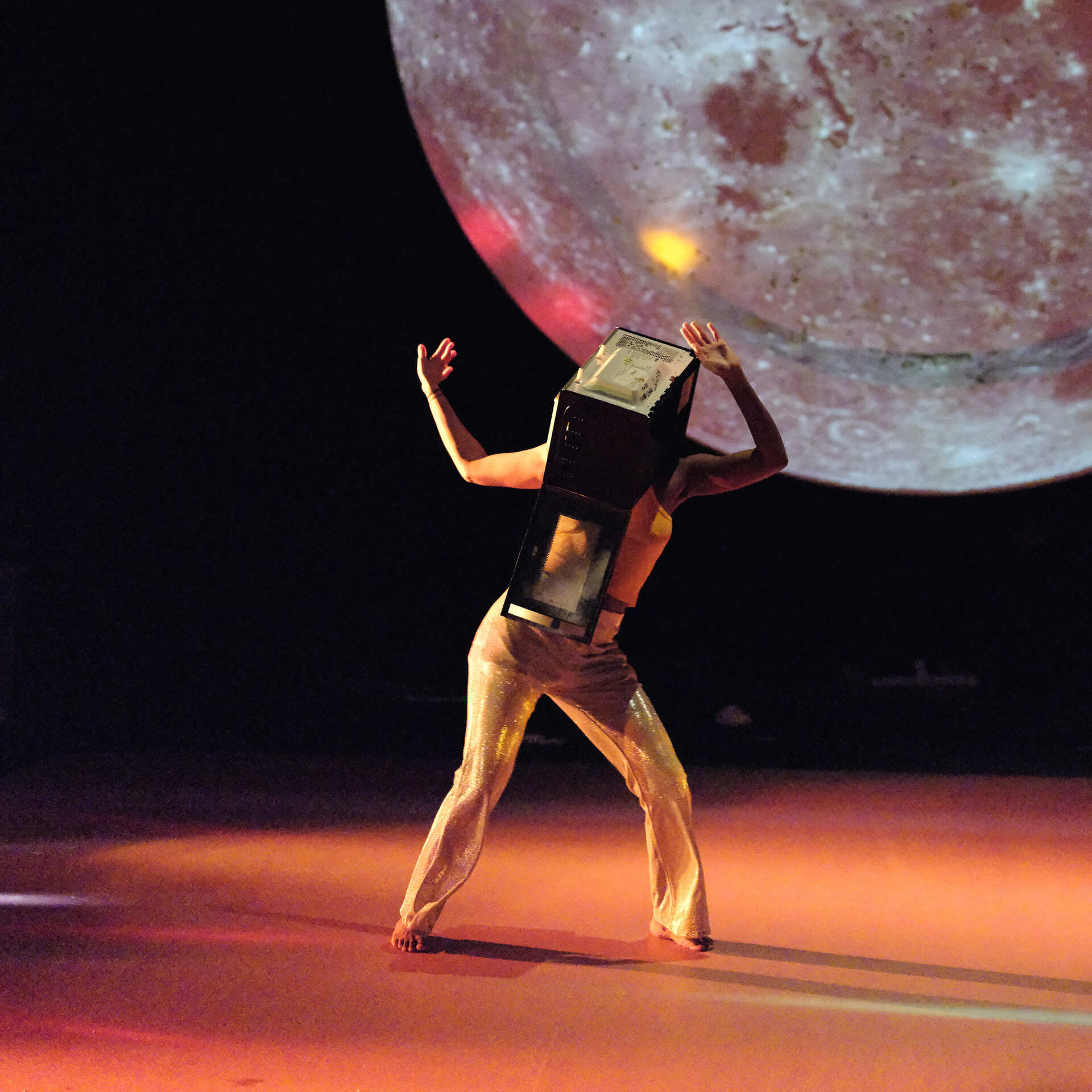 Sur une scène éclairée en orange, une personne danse, les bras levés. Sa tête est entièrement recouverte par un ancien téléviseur cubique. En arrière-plan, une grande image de la lune est projetée sur un écran.