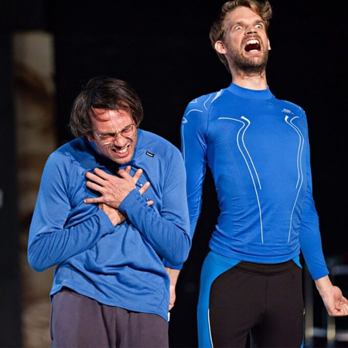 Deux comédiens sur scène portant des hauts de sport bleus identiques. L'un est courbé, grimaçant et serrant sa poitrine dans une expression de douleur feinte, tandis que l'autre, debout, hurle vers le ciel avec une expression faciale exagérée et comique.