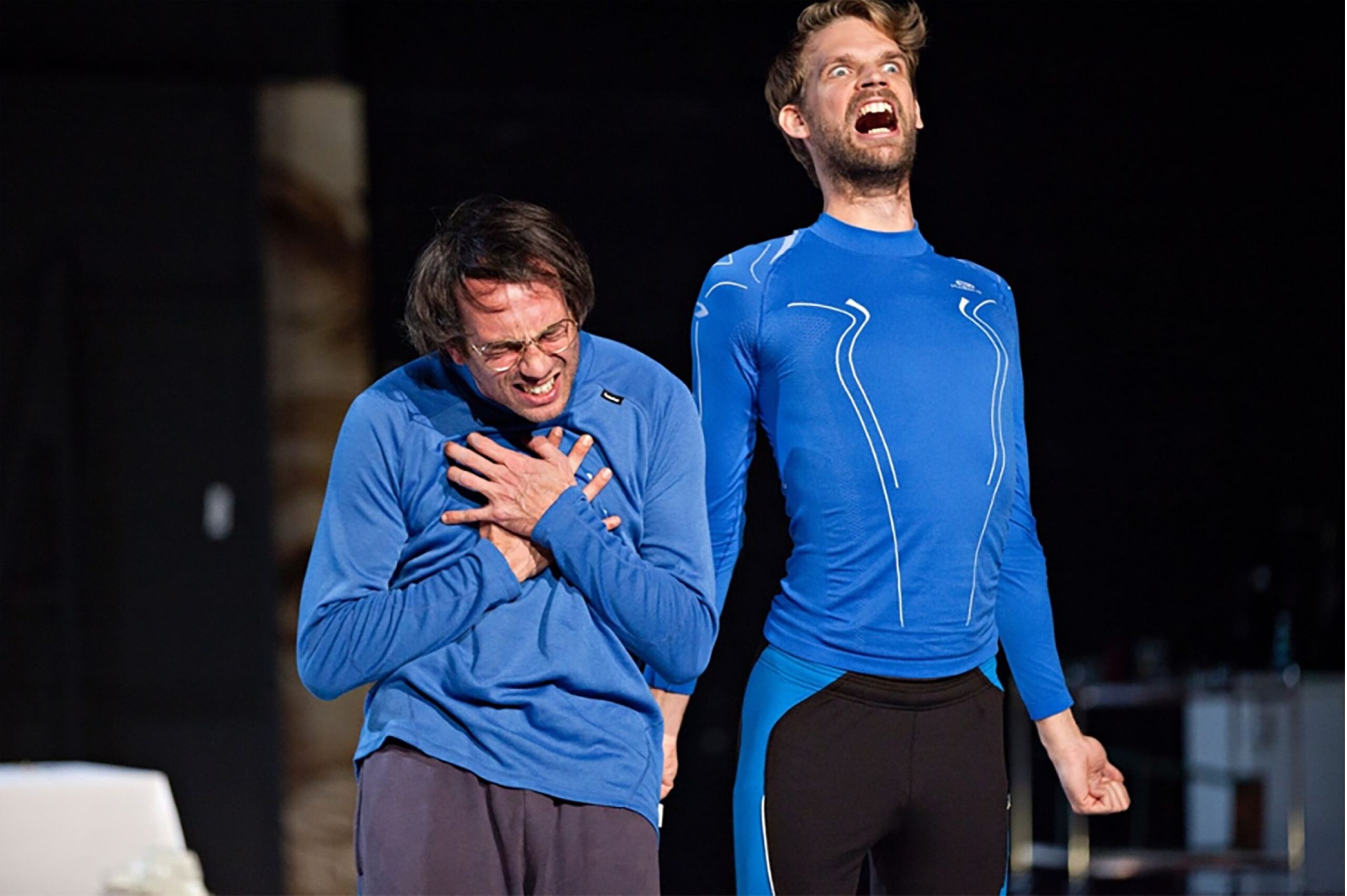 Deux comédiens sur scène portant des hauts de sport bleus identiques. L'un est courbé, grimaçant et serrant sa poitrine dans une expression de douleur feinte, tandis que l'autre, debout, hurle vers le ciel avec une expression faciale exagérée et comique.