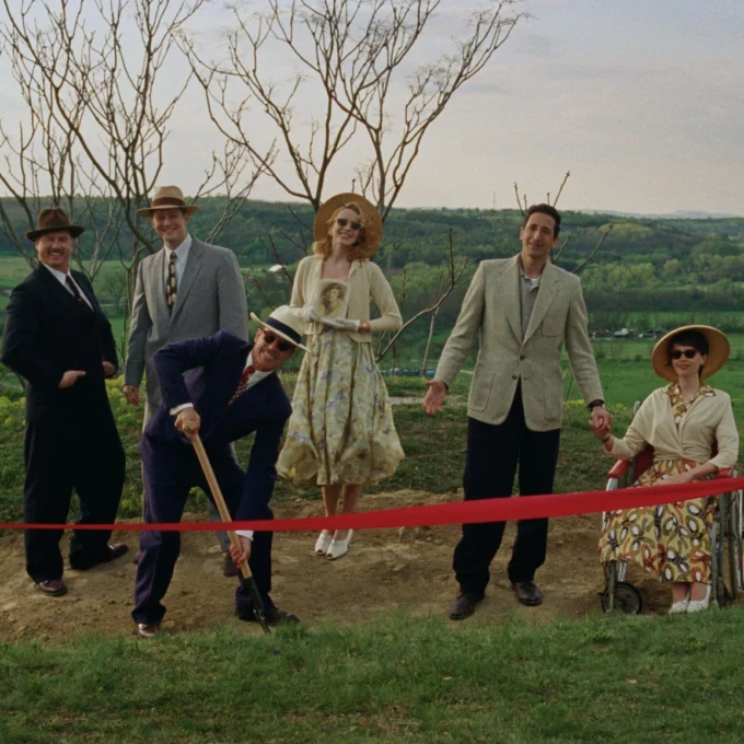Une image du film 'The Brutalist' montrant un groupe de personnes en tenues d'époque sur une colline pour une cérémonie d'inauguration, avec un homme creusant le sol devant un ruban rouge.