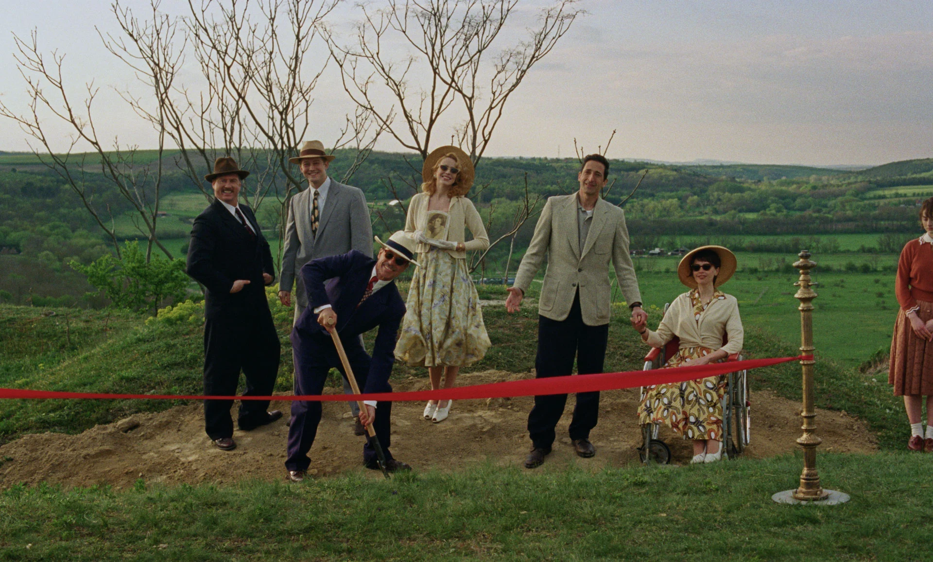 Une image du film 'The Brutalist' montrant un groupe de personnes en tenues d'époque sur une colline pour une cérémonie d'inauguration, avec un homme creusant le sol devant un ruban rouge.