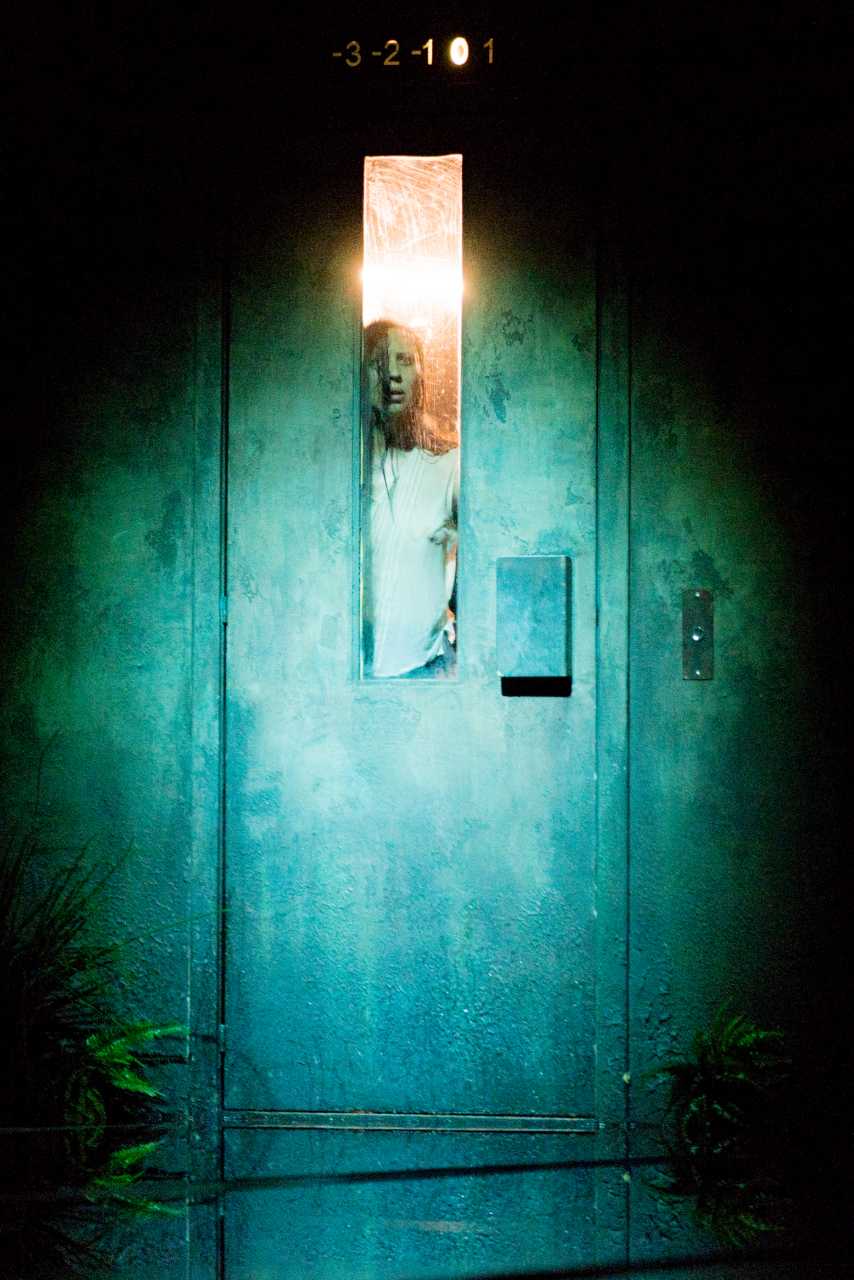 On aperçoit un homme derrière la vitre rectangulaire d'une porte d'ascenceur. Il est debout et la porte est éclairé par un spot.