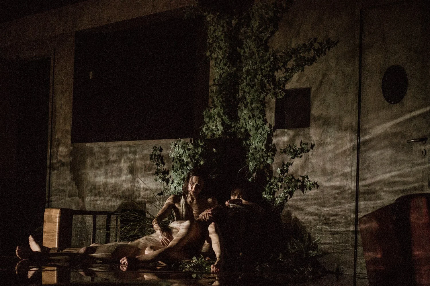 Un danseuse soutient un danseur, ils sont allongés dans l'eau au milieu d'un décors très sombre et végétalisé.