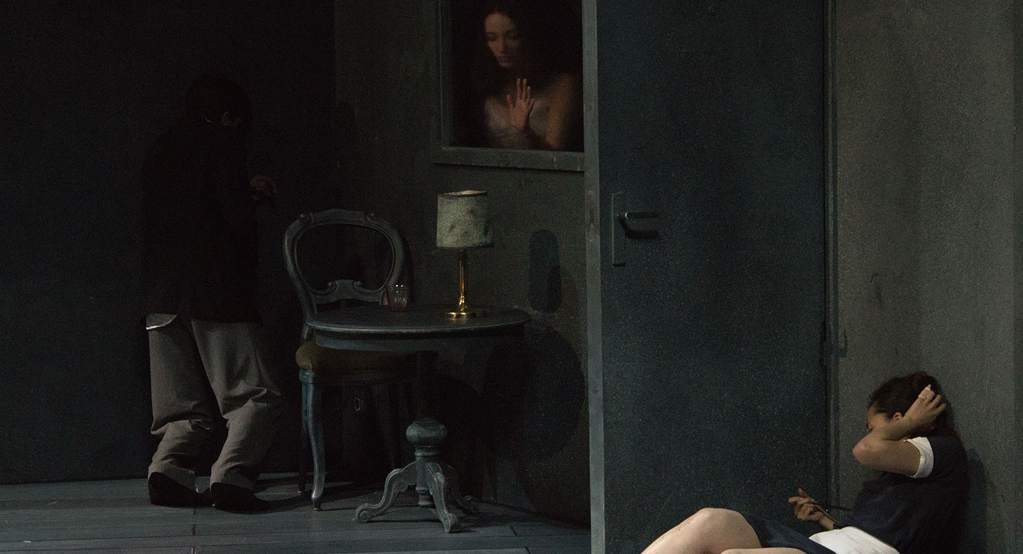 Un danseuse habillée en femme de ménage est adossé à un mur, on dirait se soutient la tête comme si elle s'était fait mal. Par une fenêtre, on aperçoit le buste d'une danseuse, l'encadrement de la fenêtre fait penser à un tableau figé.