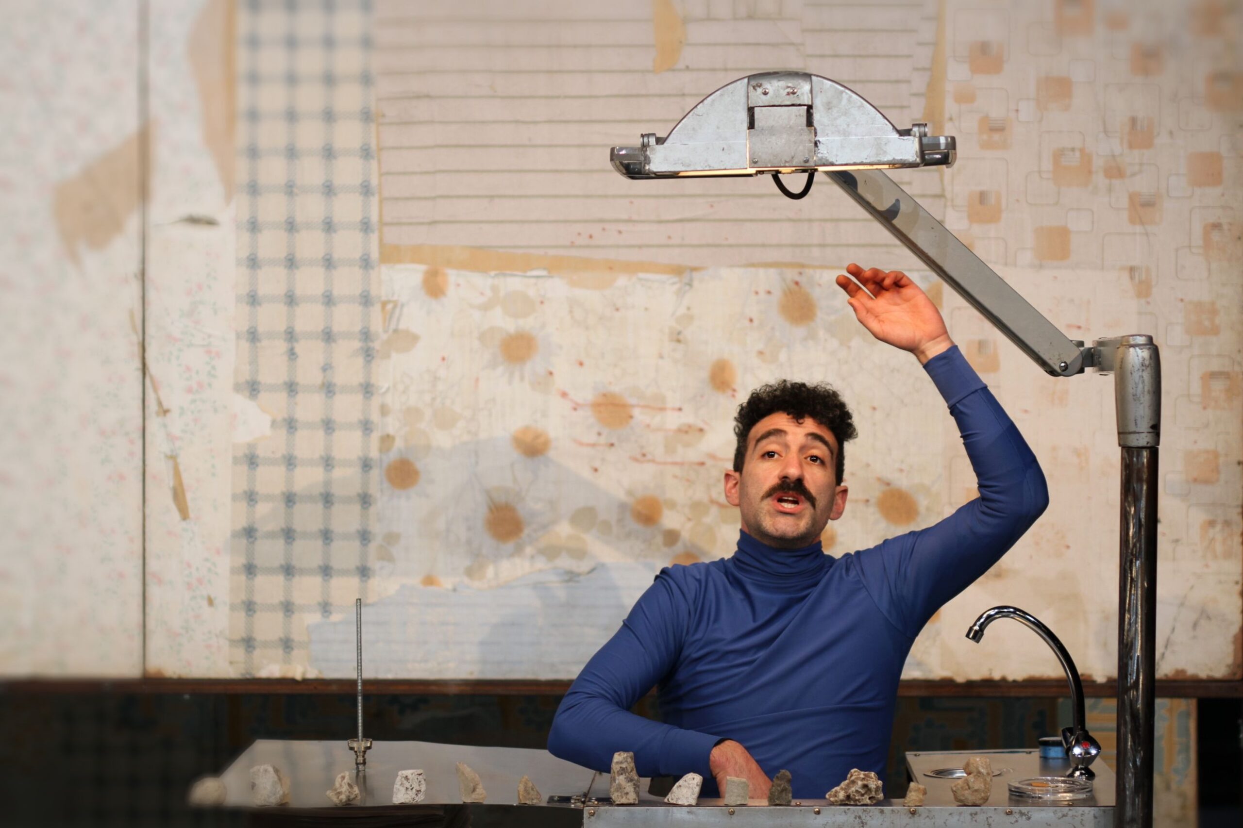 Plan moyen d'un homme moustachu portant un col roulé bleu, le bras levé vers une lampe industrielle articulée suspendue au-dessus de sa tête. Il se tient dans un décor au papier peint défraîchi, avec un robinet et une rangée de petites pierres posées sur une table au premier plan.