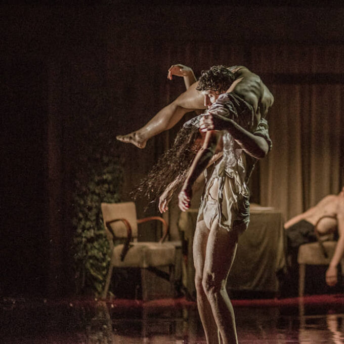 Au premier plan un couple de danseurs fait un porté, ils sont en sous-vêtement. En arrière plan, on voit un salon dans lequel une femme masse la tête d'un danseur torse nu.