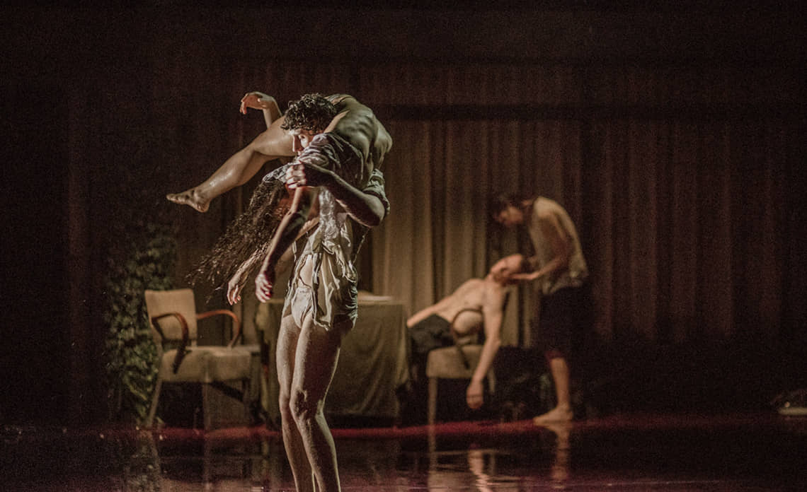 Au premier plan un couple de danseurs fait un porté, ils sont en sous-vêtement. En arrière plan, on voit un salon dans lequel une femme masse la tête d'un danseur torse nu.