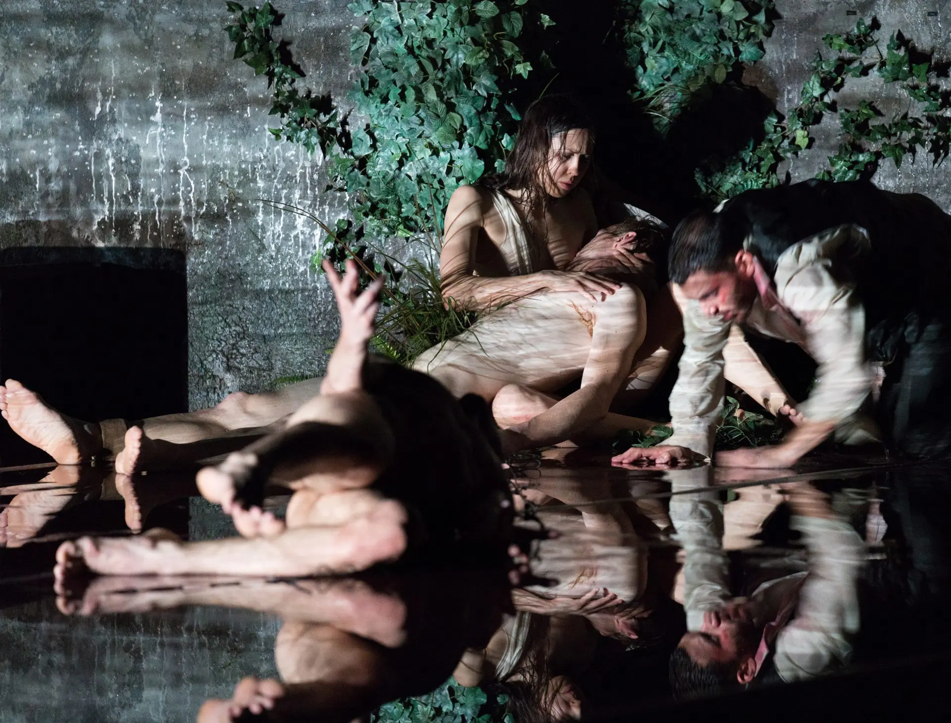 Au premier plan, une danseuse est allongée au sol en grand écart, tendant la main. En arrière-plan, soutient la tête d'un homme qui semble évanoui, créant une atmosphère de chaos et de drame. Le sol est recouvert d'eau.
