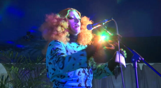 L'artiste Blanket la Goulue sur scène lors du spectacle Playback Tour, portant une volumineuse perruque rousse frisée et une robe bleue à motifs noirs, chantant au micro sous des éclairages de scène colorés et un éclat lumineux intense.