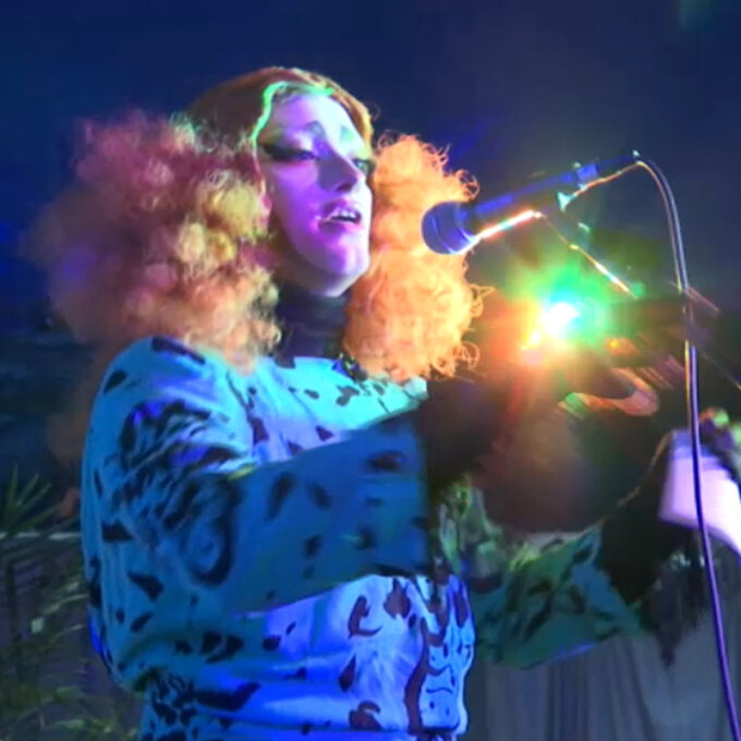 L'artiste Blanket la Goulue sur scène lors du spectacle Playback Tour, portant une volumineuse perruque rousse frisée et une robe bleue à motifs noirs, chantant au micro sous des éclairages de scène colorés et un éclat lumineux intense.