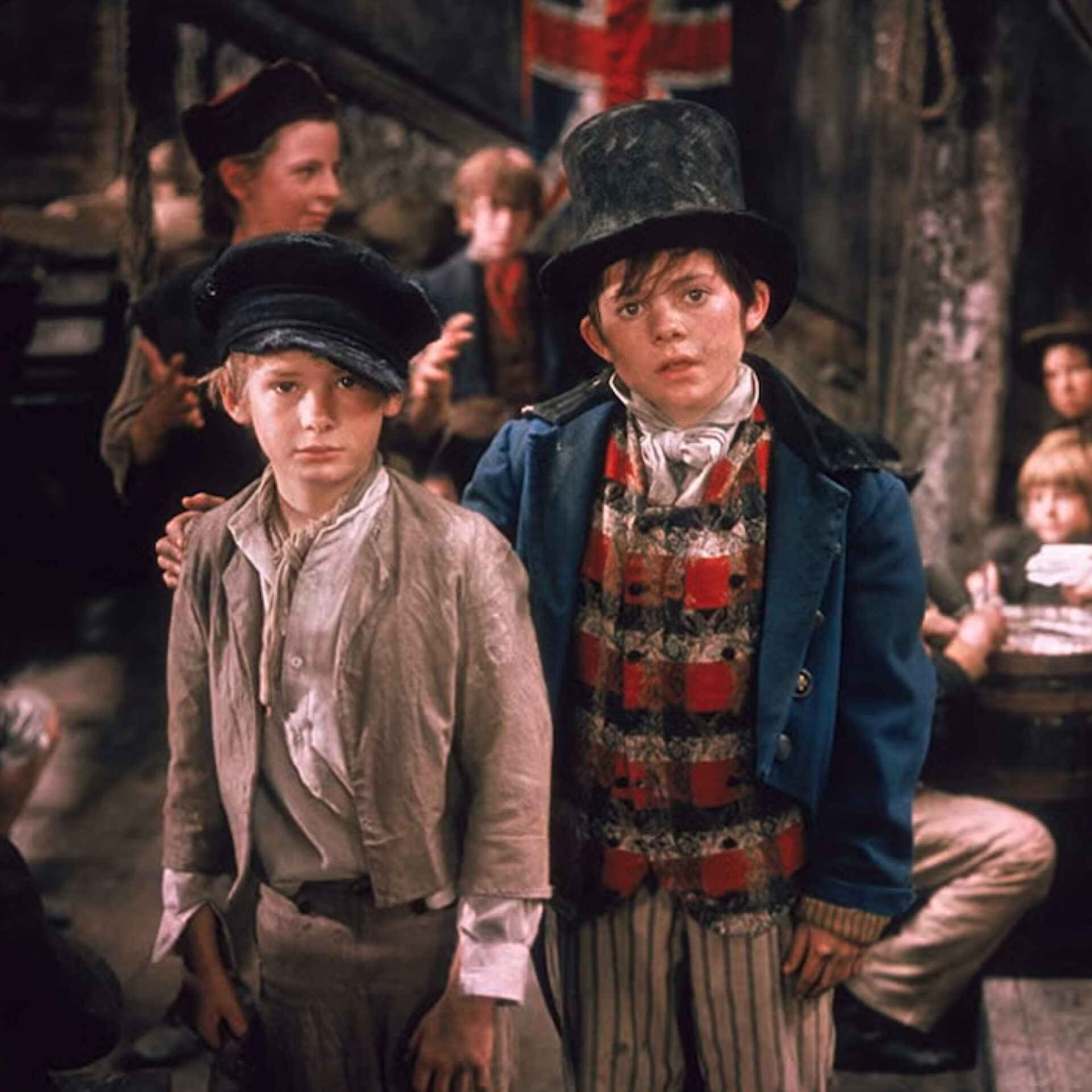 Le personnage de l'Artful Dodger (le Renard), coiffé d'un haut-de-forme, se tient à côté du jeune Oliver Twist dans un décor victorien bondé, tiré du film 