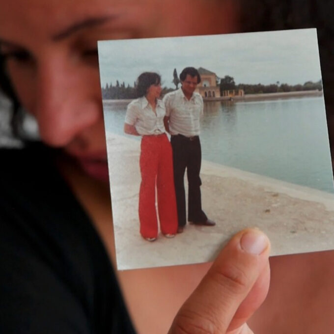 Gros plan sur une main tenant une petite photo carrée vintage montrant un homme et une femme (en pantalon rouge) posant devant un plan d'eau au Maroc. En arrière-plan, le visage flou d'une femme observe la photo avec émotion. Visuel pour le spectacle 