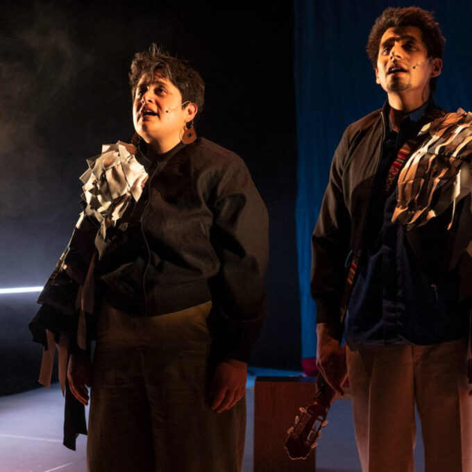 Plan moyen de deux interprètes sur une scène sombre, éclairés de façon dramatique. À gauche, une femme aux cheveux courts, et à droite, un homme tenant le manche d'une guitare. Ils regardent tous deux vers le haut avec émotion, la bouche entrouverte. Leurs vestes sombres sont ornées de lambeaux de tissu ou de papier froissé accrochés aux épaules.