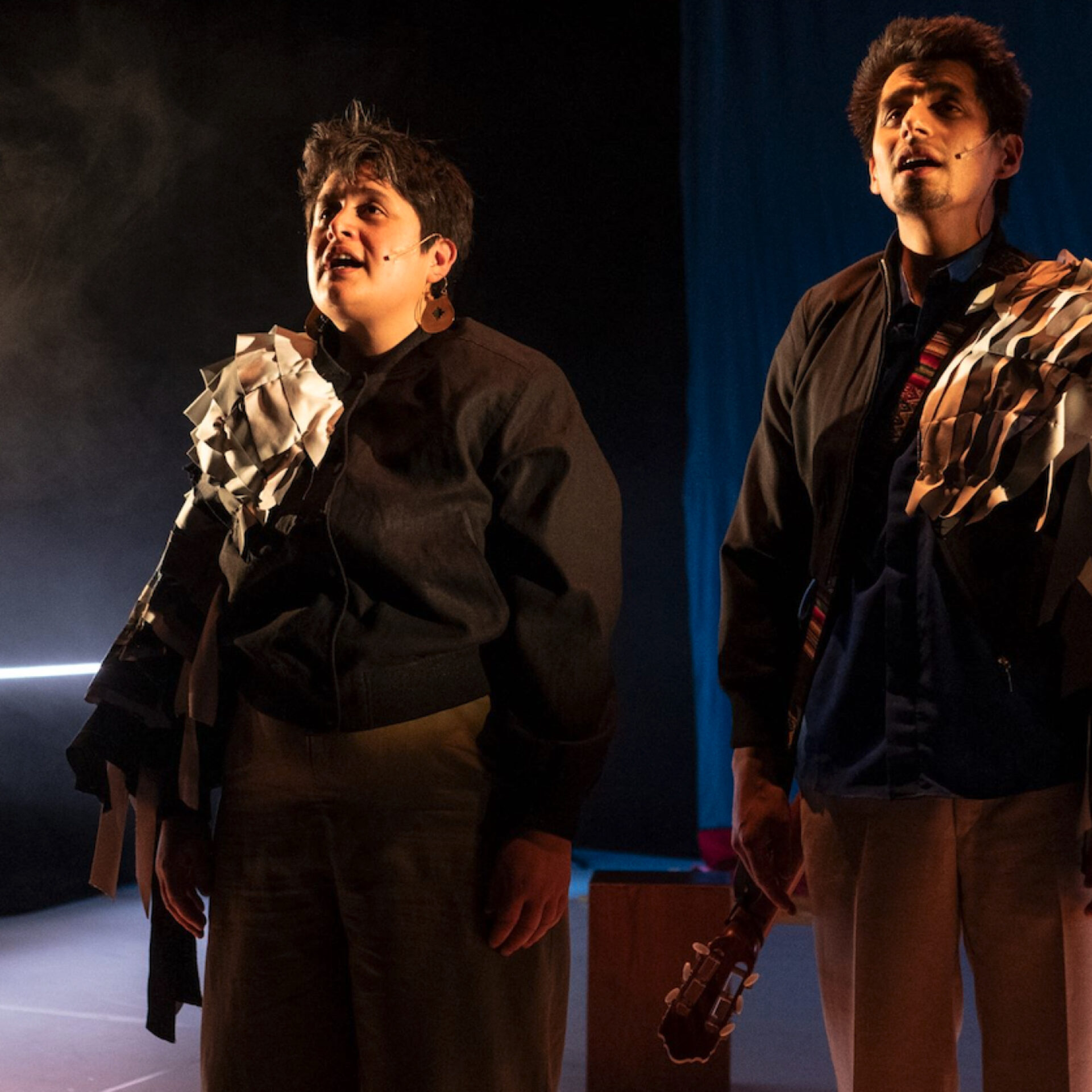 Plan moyen de deux interprètes sur une scène sombre, éclairés de façon dramatique. À gauche, une femme aux cheveux courts, et à droite, un homme tenant le manche d'une guitare. Ils regardent tous deux vers le haut avec émotion, la bouche entrouverte. Leurs vestes sombres sont ornées de lambeaux de tissu ou de papier froissé accrochés aux épaules.