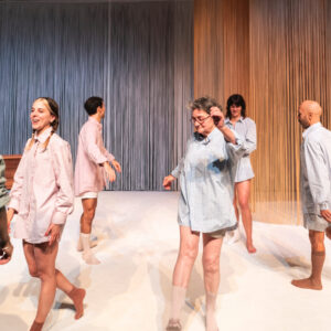 Photo de scène lumineuse montrant un groupe de cinq comédiens de différents âges, vêtus uniquement de grandes chemises pastel (roses et bleues) et de chaussettes beiges. Ils déambulent ou dansent avec le sourire sur un sol blanc, devant un fond composé de longs fils verticaux éclairés en bleu à gauche et en orange à droite.