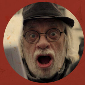 Portrait en gros plan d'un homme âgé avec une barbe blanche, des lunettes rondes et un chapeau sombre. Il a la bouche grande ouverte et les yeux écarquillés dans une expression de cri ou de surprise intense.