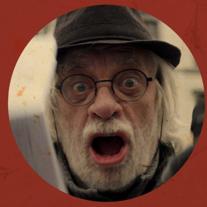 Portrait en gros plan d'un homme âgé avec une barbe blanche, des lunettes rondes et un chapeau sombre. Il a la bouche grande ouverte et les yeux écarquillés dans une expression de cri ou de surprise intense.