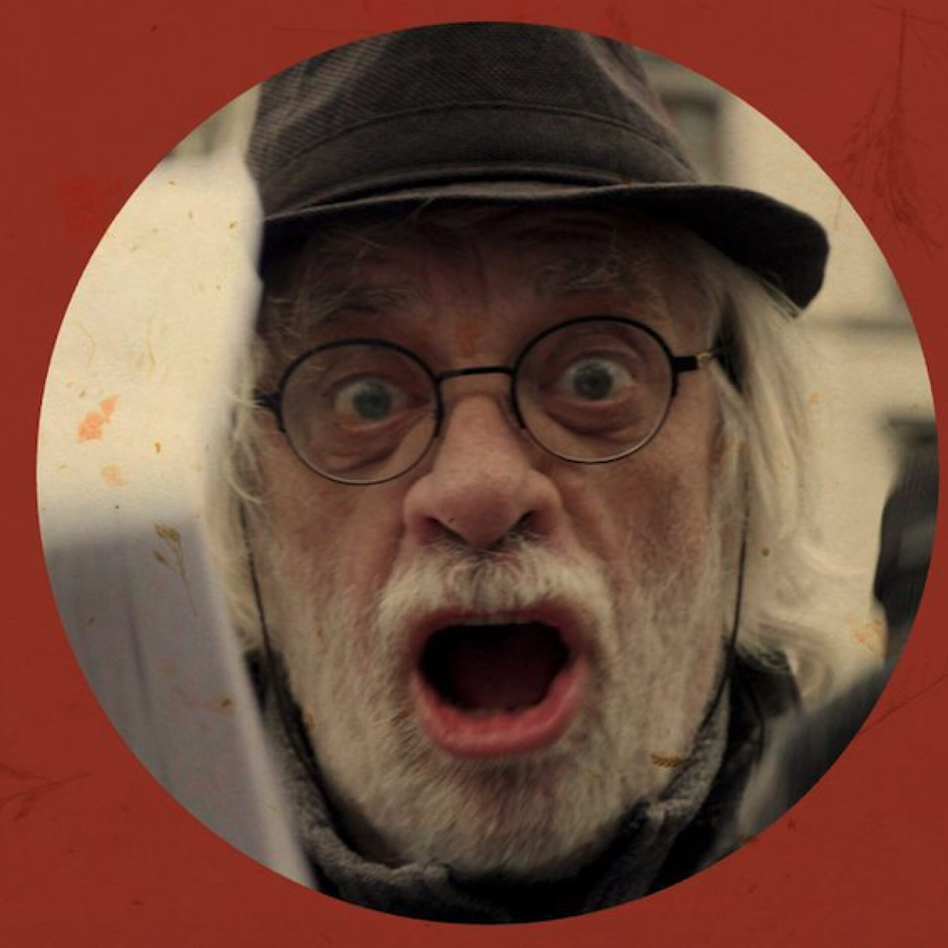 Portrait en gros plan d'un homme âgé avec une barbe blanche, des lunettes rondes et un chapeau sombre. Il a la bouche grande ouverte et les yeux écarquillés dans une expression de cri ou de surprise intense.