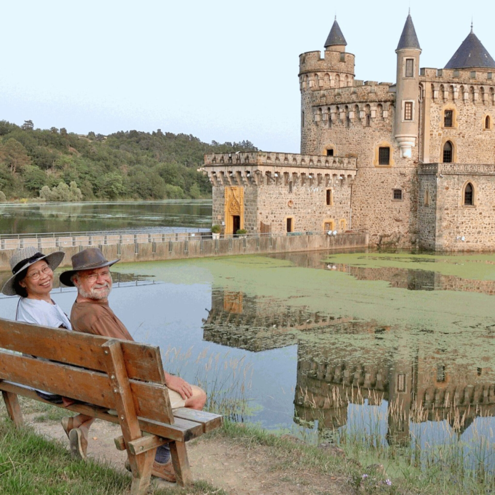 Un couple de seniors souriant est assis sur un banc en bois au premier plan. La femme, à gauche, porte un chapeau de paille clair et un haut blanc. L'homme, à droite, a une barbe grise et porte un chapeau de type fedora. En arrière-plan, un magnifique château médiéval en pierre avec des tours et des créneaux se dresse au bord d'un lac ou de douves. Le reflet du château est visible dans l'eau calme, et des collines boisées complètent le paysage à l'horizon.