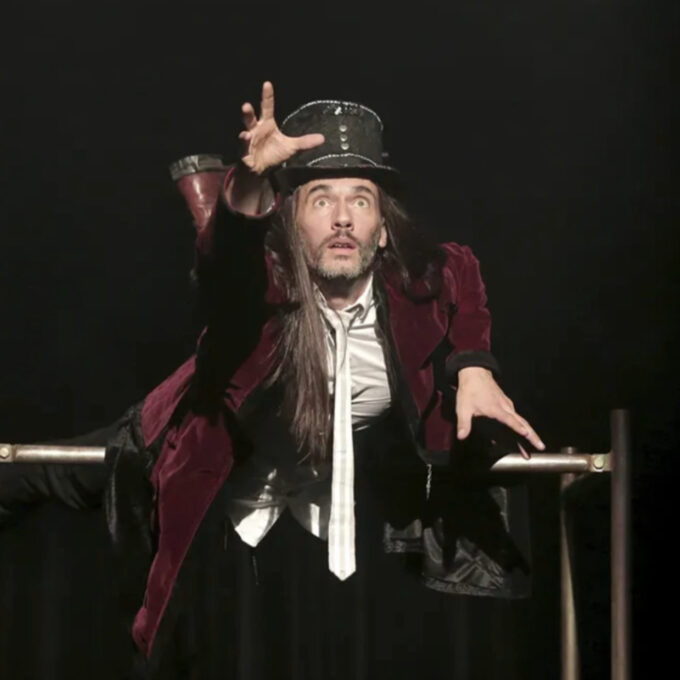 Le comédien du seul en scène Canopée, vêtu d'une veste en velours bordeaux et d'un chapeau haut de forme, est suspendu à l'horizontale sur une barre métallique, tendant la main vers l'avant avec une expression intense sur fond noir.