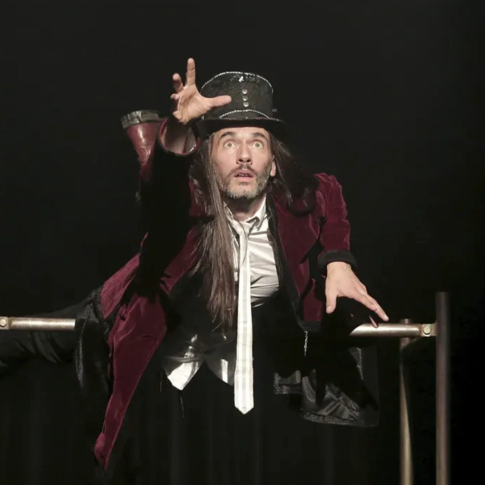 Le comédien du seul en scène Canopée, vêtu d'une veste en velours bordeaux et d'un chapeau haut de forme, est suspendu à l'horizontale sur une barre métallique, tendant la main vers l'avant avec une expression intense sur fond noir.