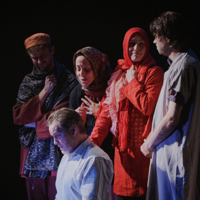 Photo de scène sur fond noir. Au premier plan, un homme en chemise blanche est assis, le regard baissé et pensif. Derrière lui, un groupe de quatre personnages (deux femmes et deux hommes) portant des vêtements et foulards traditionnels se tient debout, semblant l'observer ou s'adresser à lui avec émotion.