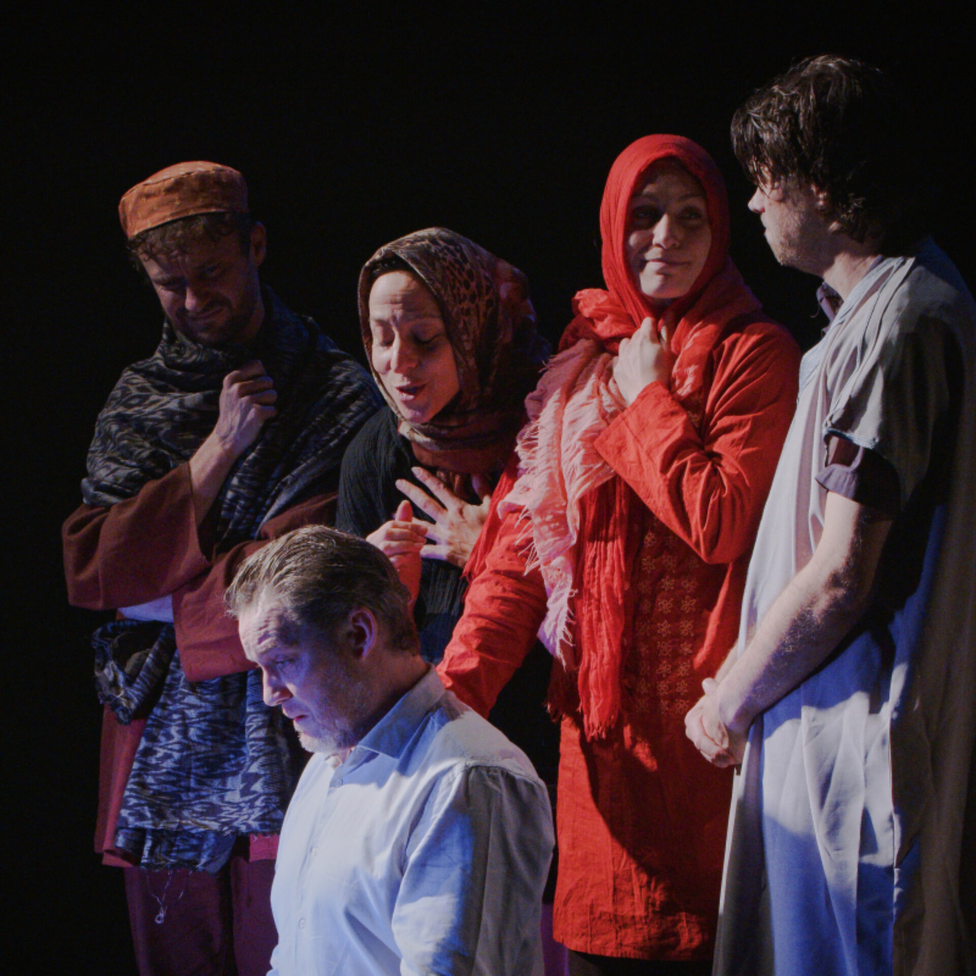 Photo de scène sur fond noir. Au premier plan, un homme en chemise blanche est assis, le regard baissé et pensif. Derrière lui, un groupe de quatre personnages (deux femmes et deux hommes) portant des vêtements et foulards traditionnels se tient debout, semblant l'observer ou s'adresser à lui avec émotion.