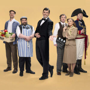 Photo de groupe de six comédiens de la pièce 'Le Schpountz'. Au centre, un homme en smoking noir pose fièrement. Il est entouré de personnages aux costumes variés : un homme portant une cagette de légumes, un autre en tablier rayé, un homme tenant un clap de cinéma, une femme en tailleur d'époque et un homme déguisé en Napoléon.