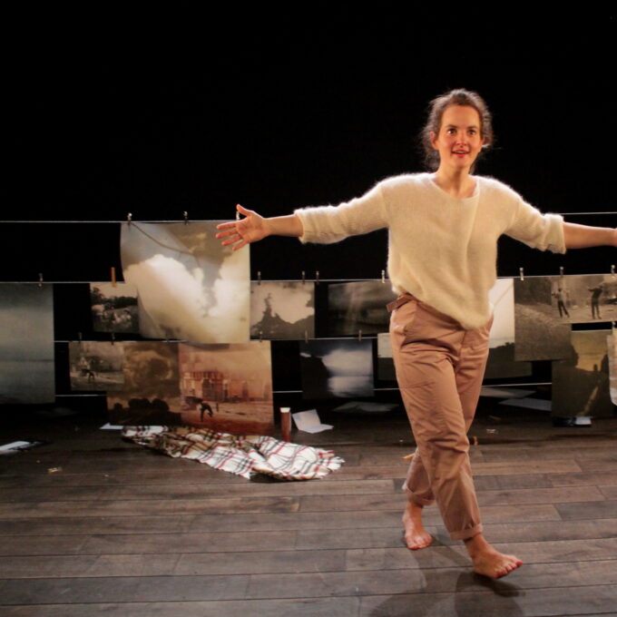 Kelly Rivière seule scène, debout les bras écartés avec en arrière plan des impressions photos accrochés à des pinces à linges. Elles semblent représenter des images de manifestations en Irlande.