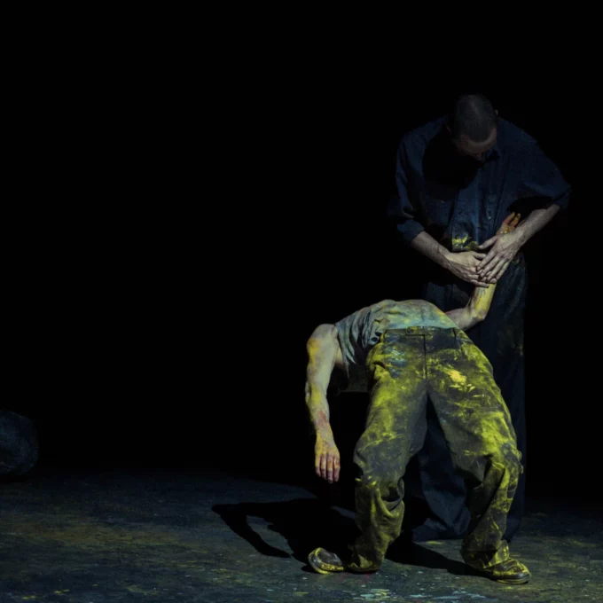 Deux danseurs, l'un est en train de faire un pont, il a la tête renversé en arrière et un pantalon taché de peinture jaune. L'autre danseur lui tient le bras, on le distingue à peine.