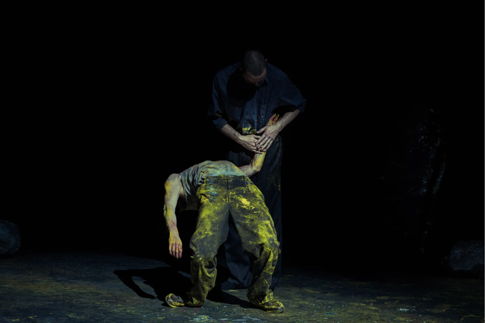 Deux danseurs, l'un est en train de faire un pont, il a la tête renversé en arrière et un pantalon taché de peinture jaune. L'autre danseur lui tient le bras, on le distingue à peine.